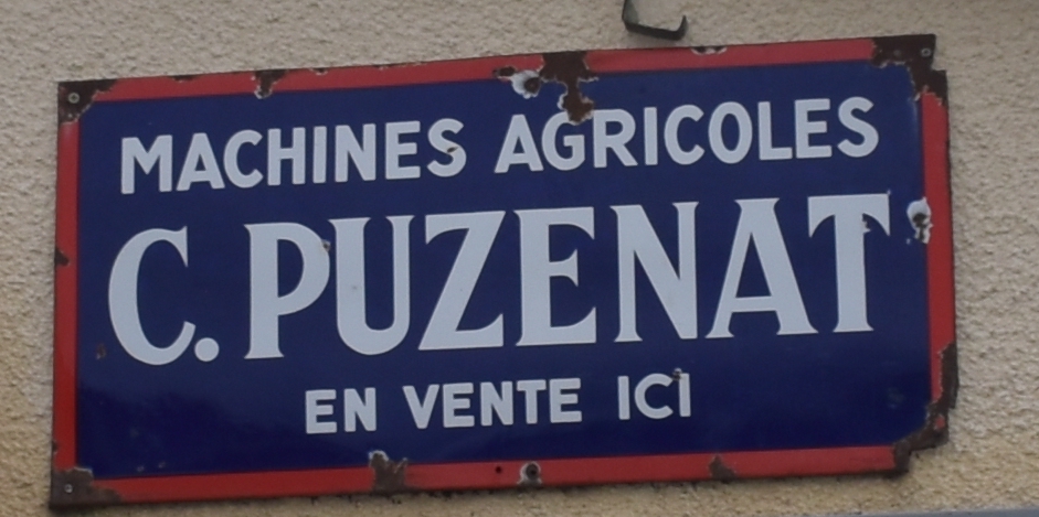 Plaque émaillée bleue à filet rouge, fixée sur un mur, portant en lettres blanches « Machines agricoles C. Puzenat — en vente ici »